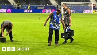 Three men hold buckets of grass on a football pitch