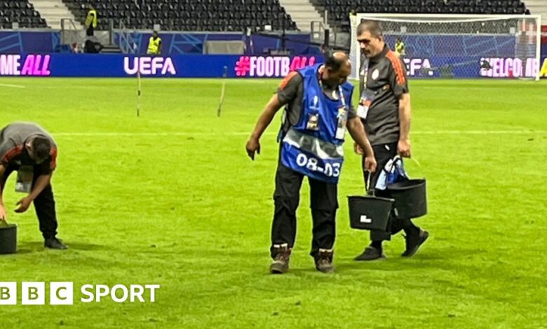 Three men hold buckets of grass on a football pitch